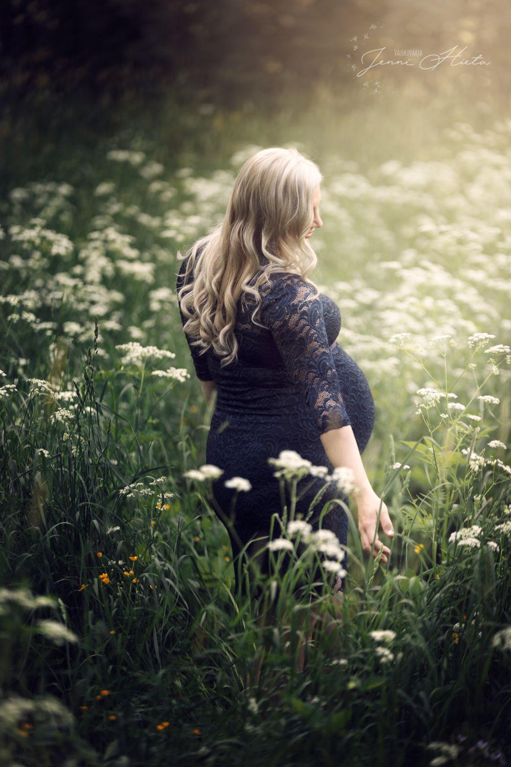 A figure in a dark lace dress stands among wild daisies in a dreamy sunlit meadow with glowing backlight.