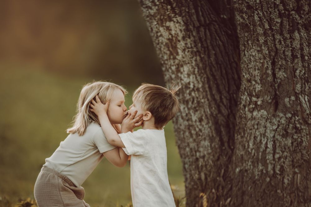 Two young children share a sweet moment while playing by a large tree trunk in soft natural lighting.