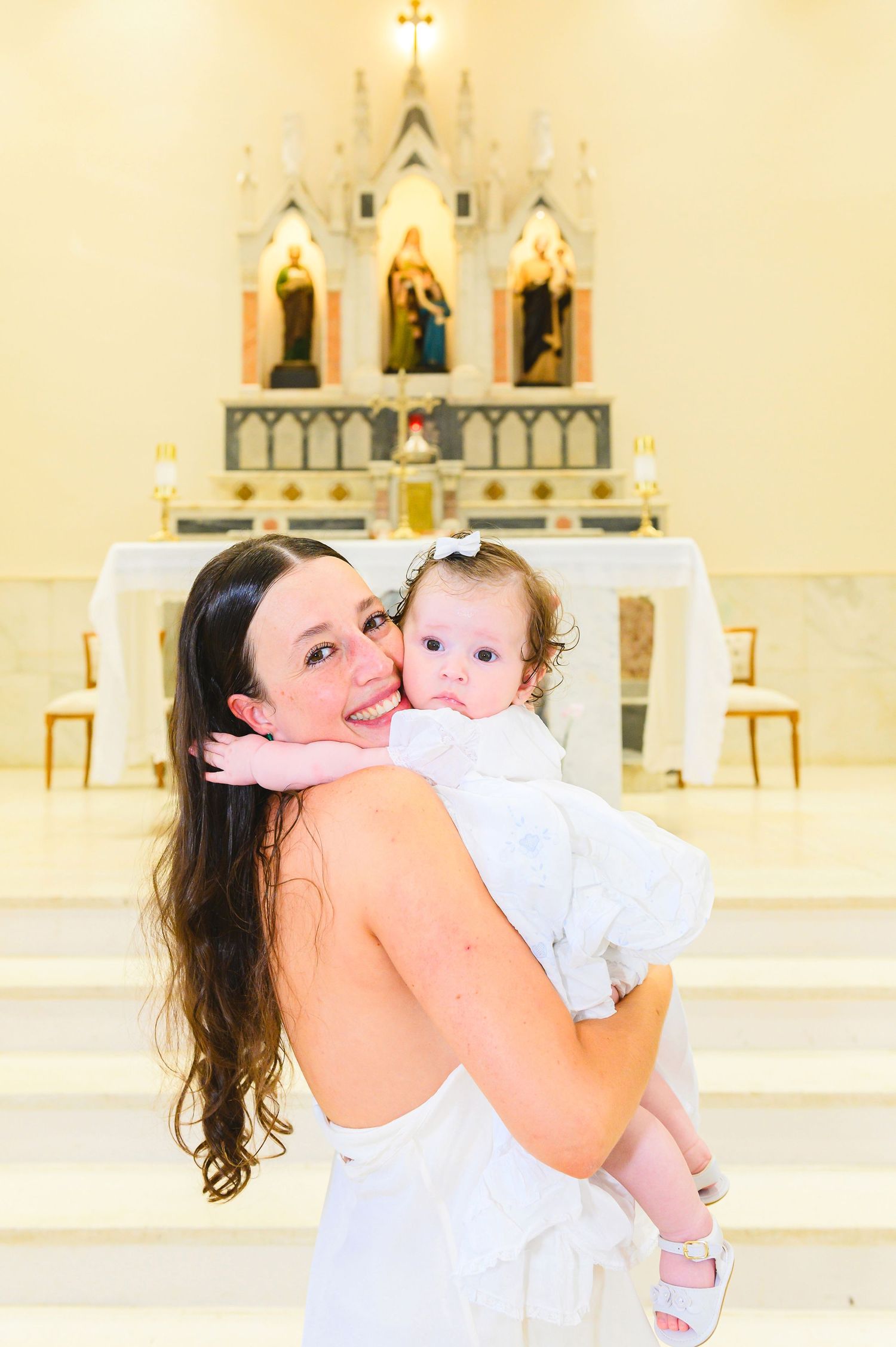 A baby in white baptismal dress being held inside a church with religious altar and statues in the background.