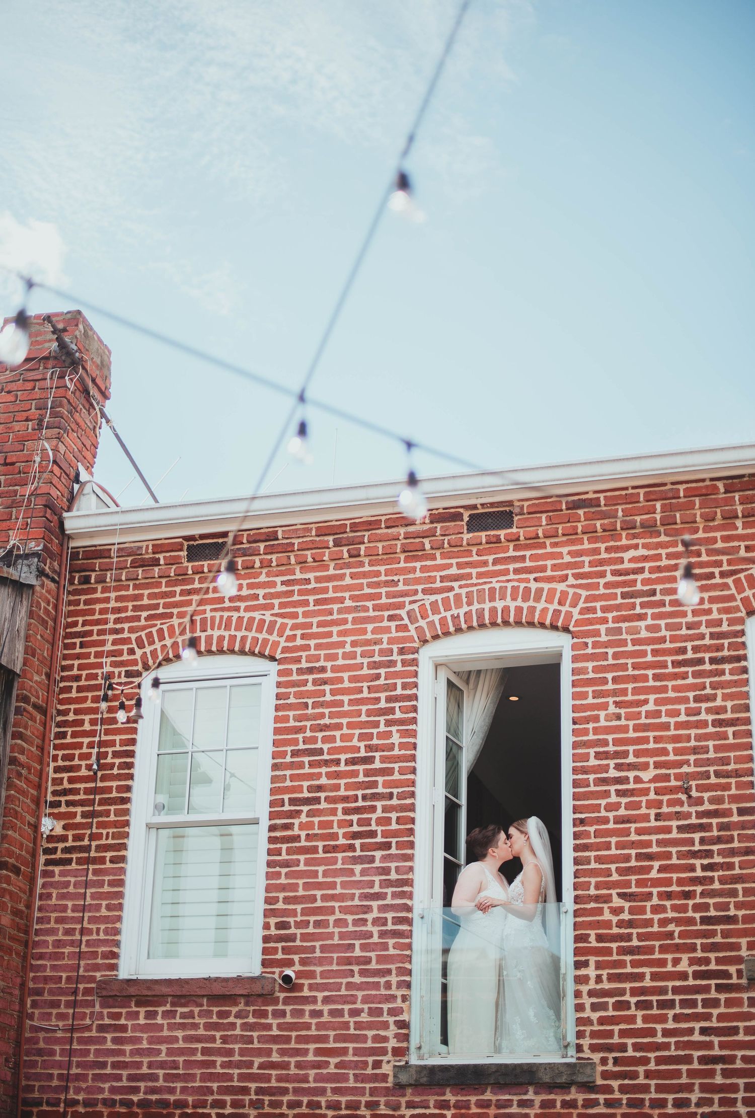 A vintage brick building with string lights overhead and a figure standing in a second floor doorway.