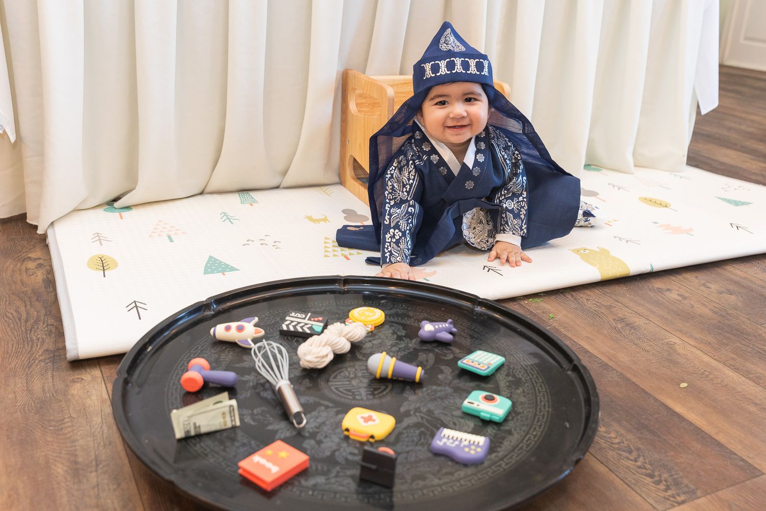 Baby in blue hoodie lying on play mat with various colorful toys scattered on round tray nearby.