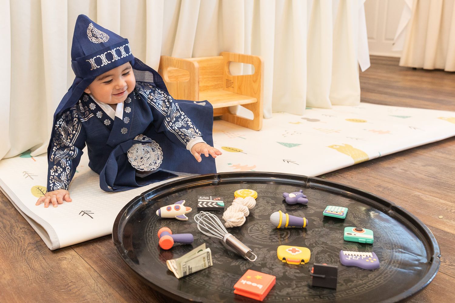 Small child wearing a blue winter hat plays with colorful toy cars on a large circular tray while looking at a book.