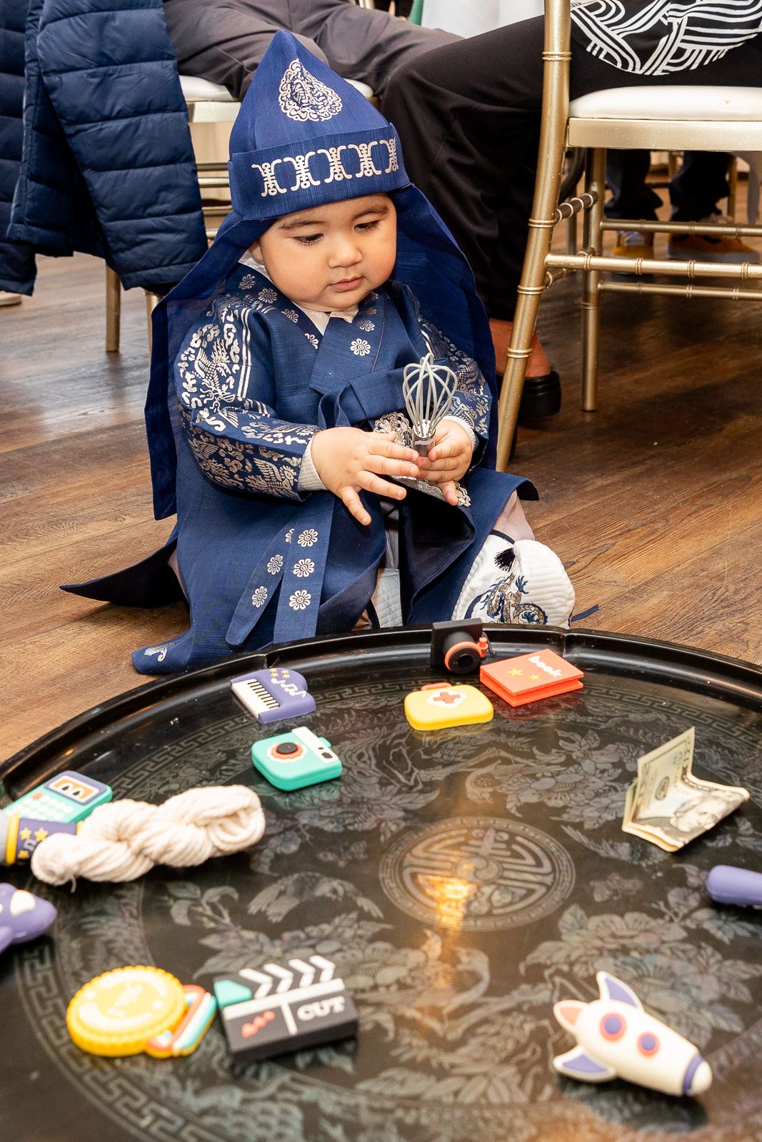 Young child in traditional blue outfit plays with colorful toys and objects on a decorative black table.