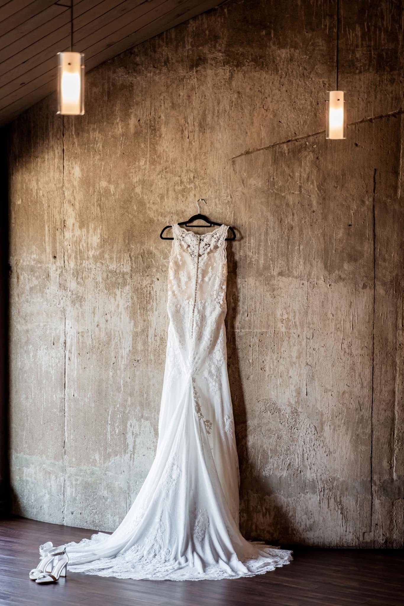 A white lace wedding dress hangs elegantly against a rustic textured wall with glowing sconce lighting above.