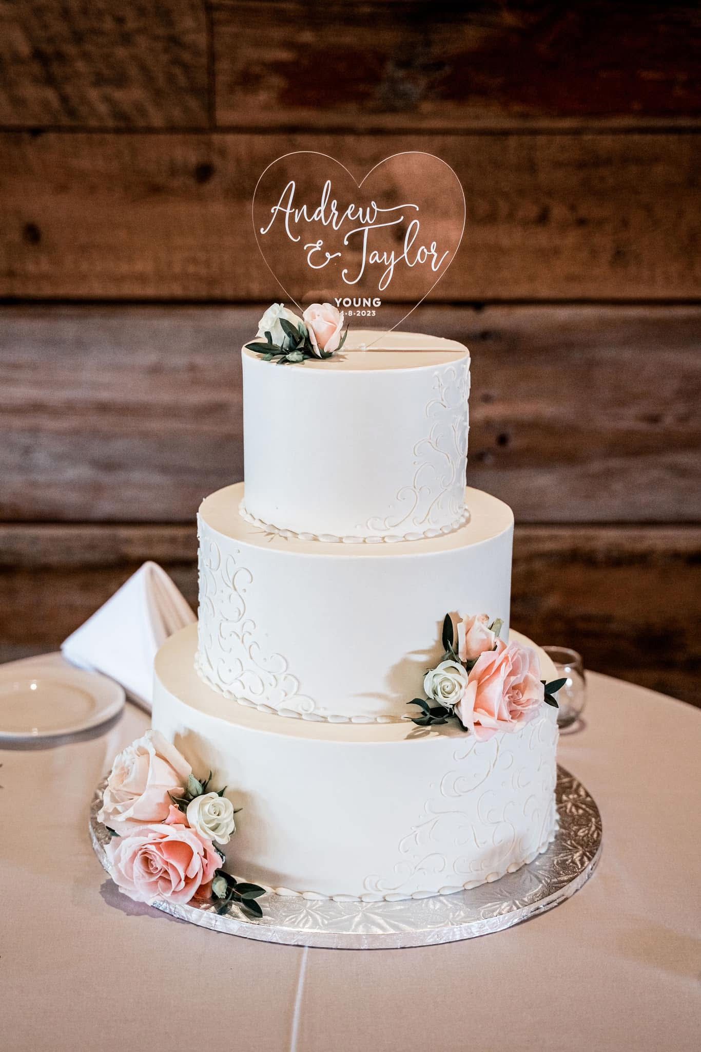 Elegant three-tier white wedding cake with lace details, pink roses, and heart-shaped acrylic topper on wooden backdrop.