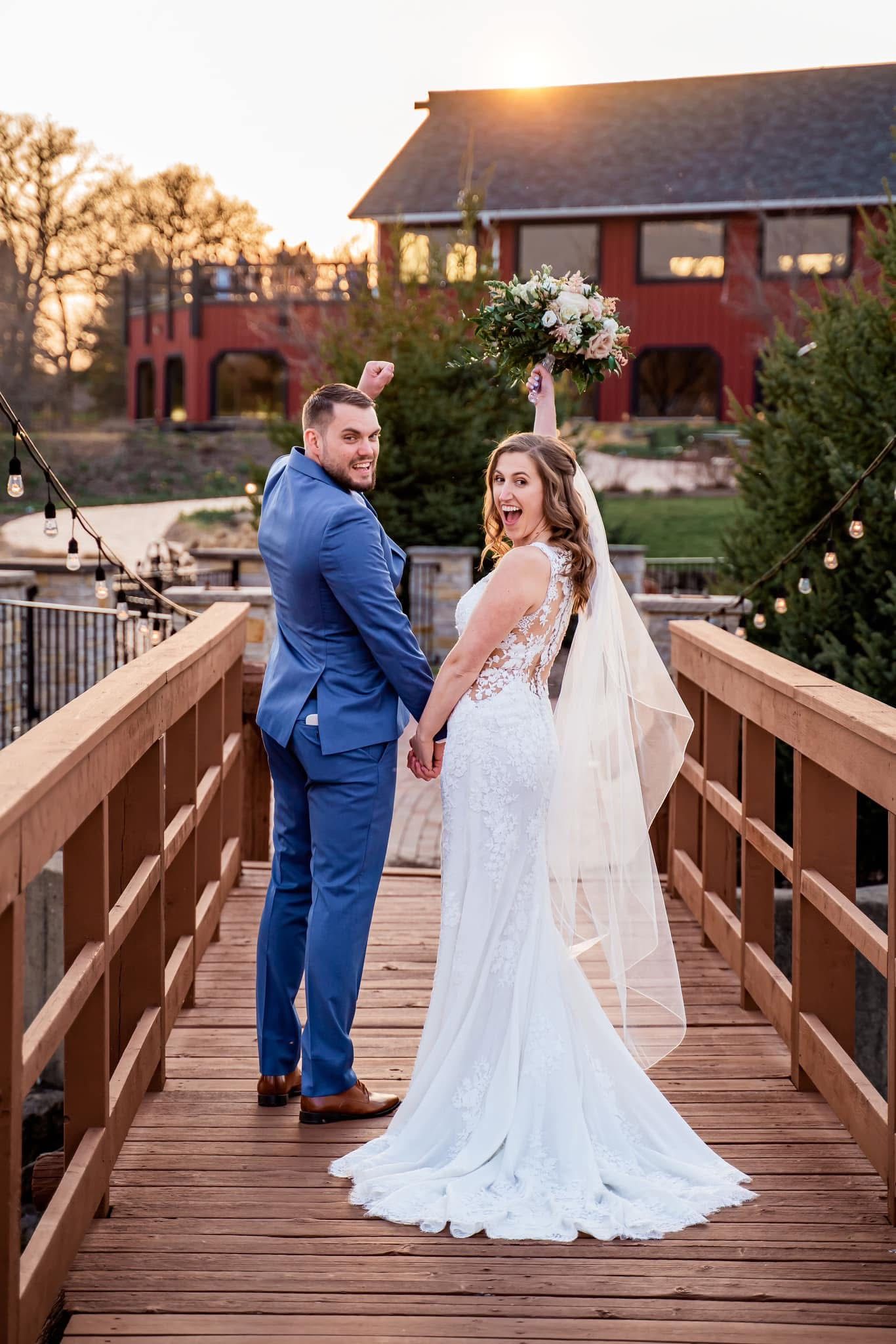 Wedding couple poses on wooden bridge at sunset with rustic red barn venue in background at golden hour.