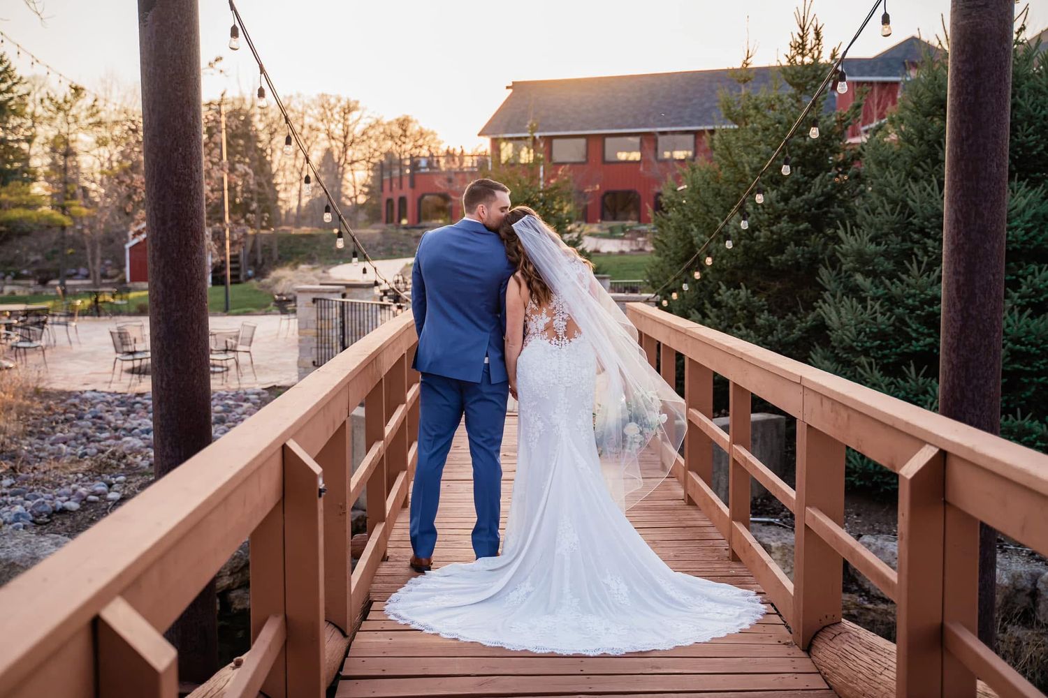 A couple in wedding attire hold hands while walking across a wooden bridge at sunset with brick buildings in background.