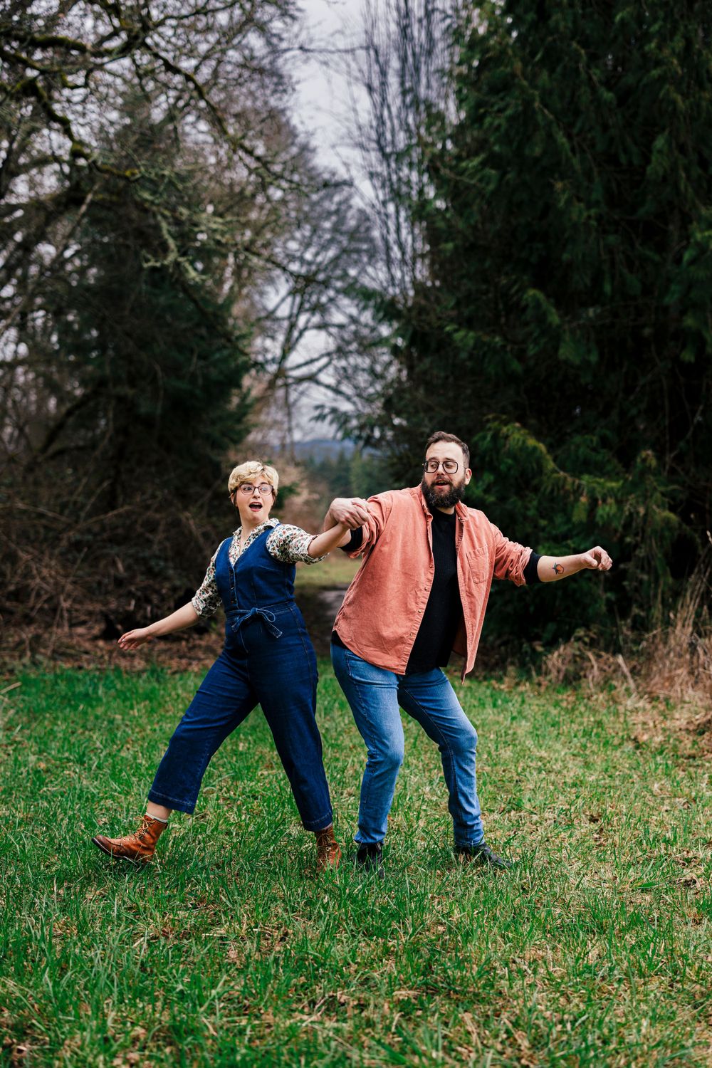 A cheerful couple strikes playful poses while standing in a grassy field surrounded by evergreen trees.