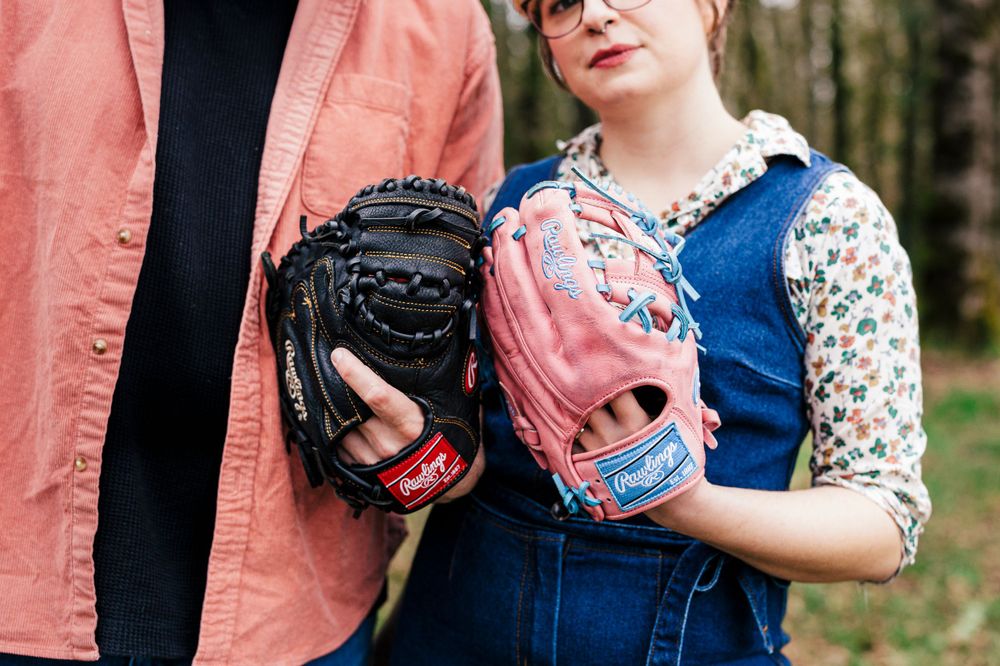 Two people share an intimate moment wearing pink and blue baseball gloves in an outdoor setting.