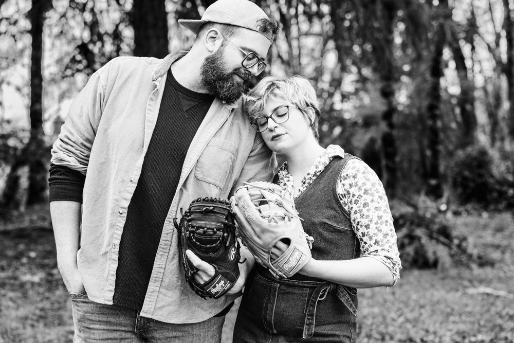 A black and white photo of a couple with baseball gloves standing together in a wooded outdoor setting.