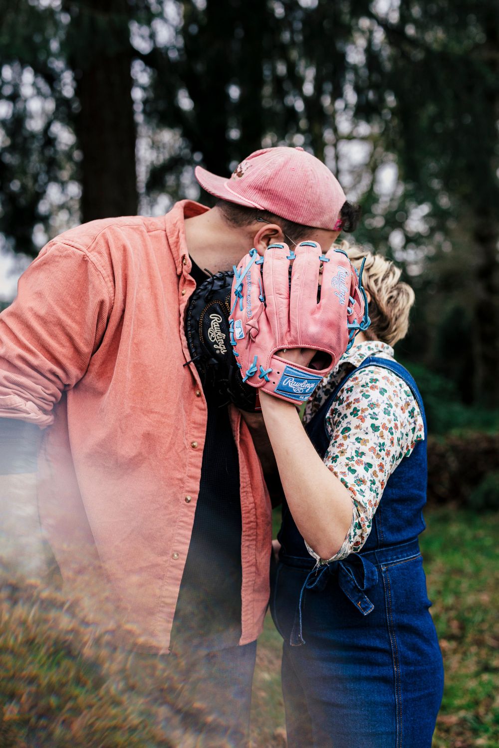 A romantic moment is captured between two people wearing matching pink baseball gloves in a forest setting.