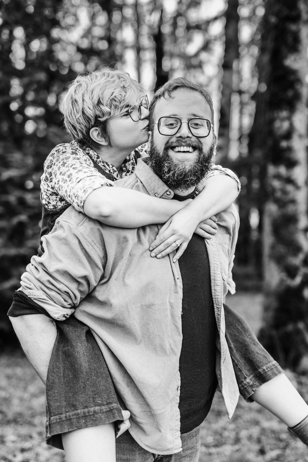 A joyful black and white photograph of two people sharing a playful piggyback ride moment in a forested setting.