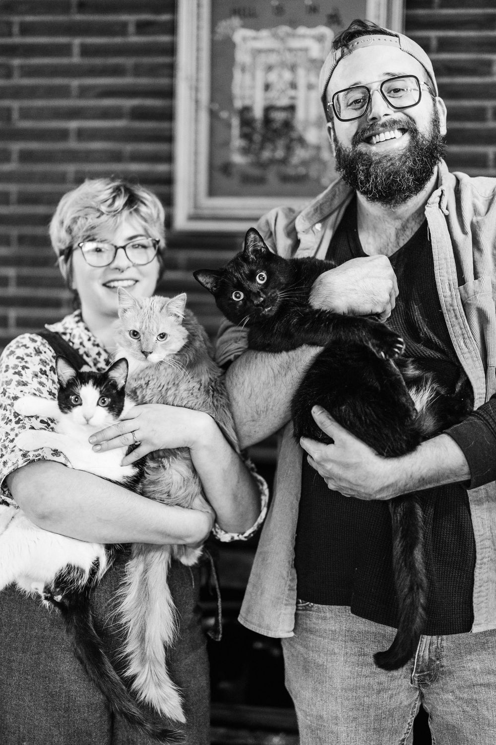 A black and white photo of pet owners holding their three cats - a black cat, a gray cat, and a calico cat.