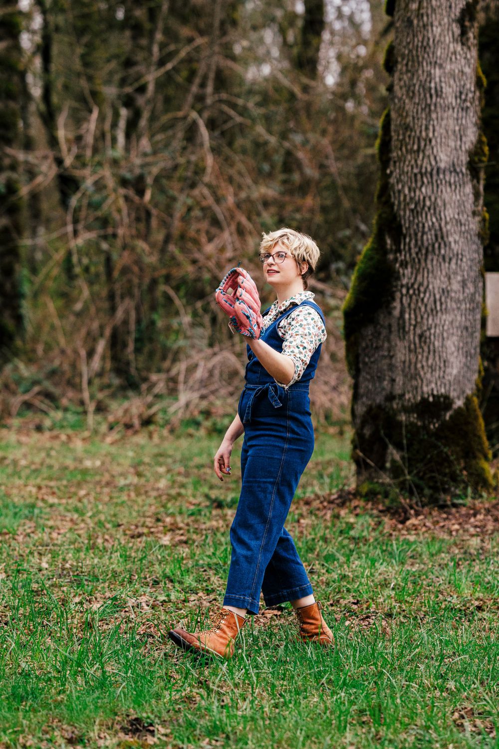 Person wearing denim overalls and a patterned sweater walking in a wooded area on a fall day.