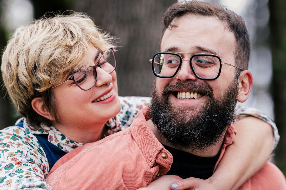 A happy couple wearing glasses embrace and smile together, with one person in a floral top and the other in a coral shirt.