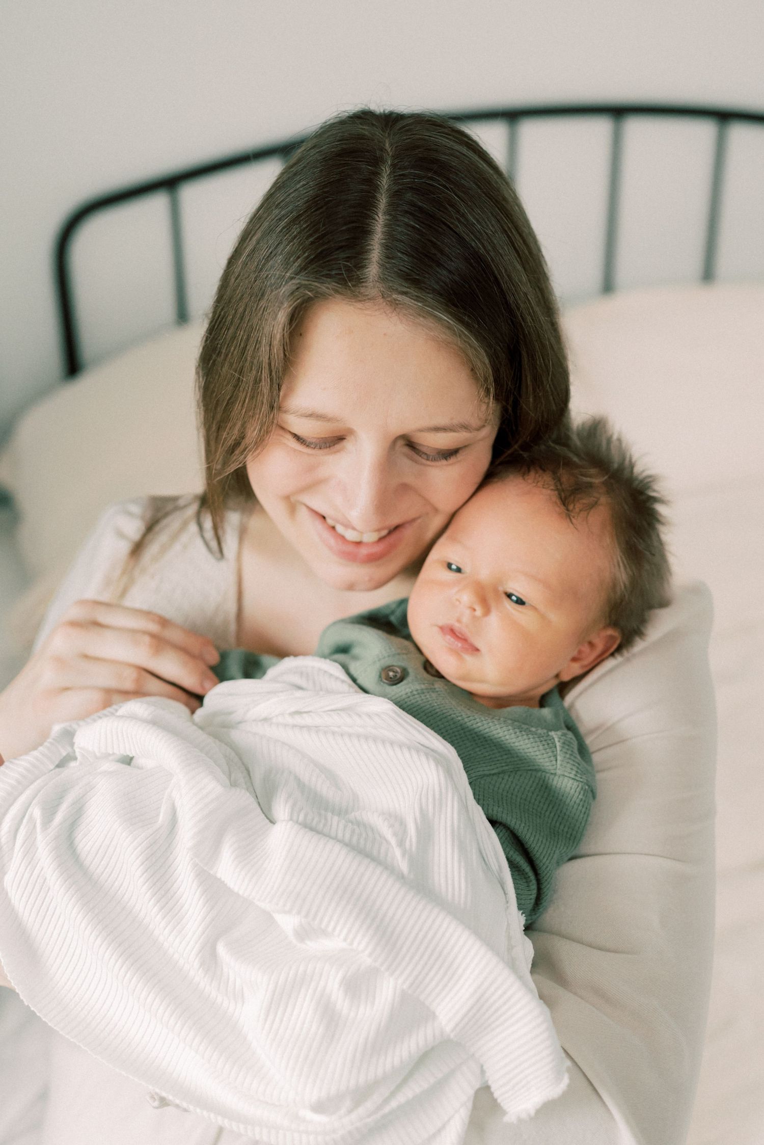 A tender moment of a mother and newborn baby snuggling together on a bed with white linens and dark headboard.