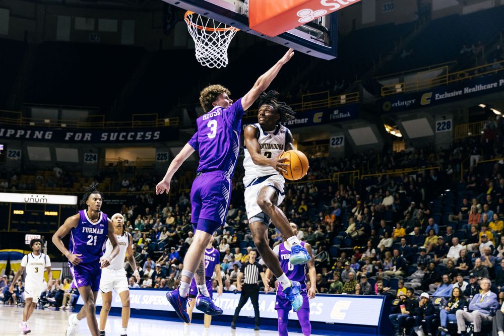 Basketball player in white jersey attempts shot while being defended by opponent in purple uniform during indoor game.