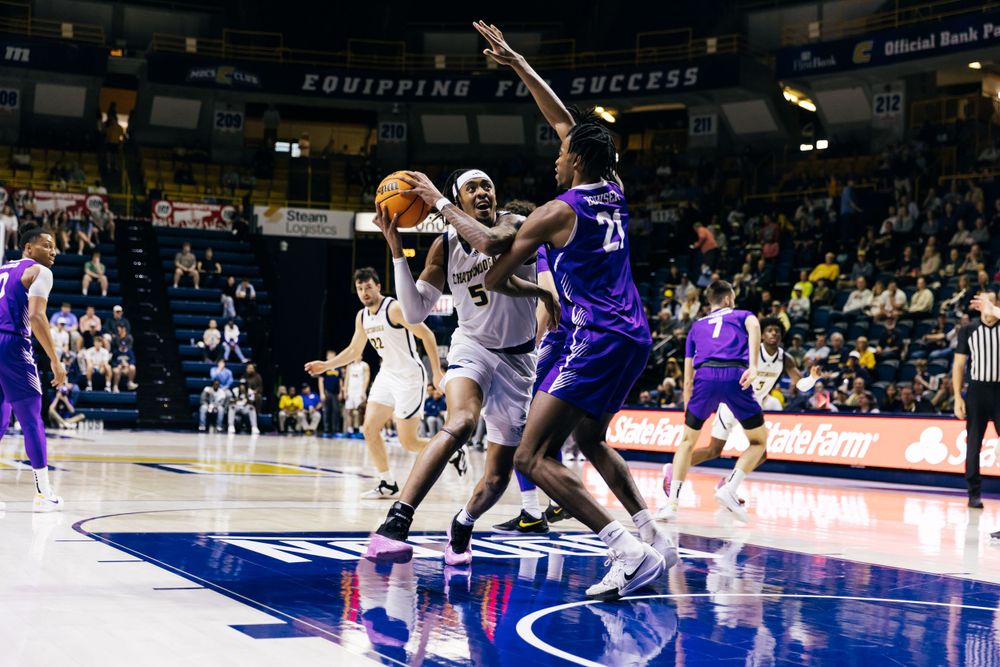 Basketball players from opposing teams compete for possession during an intense game on a brightly lit indoor court.