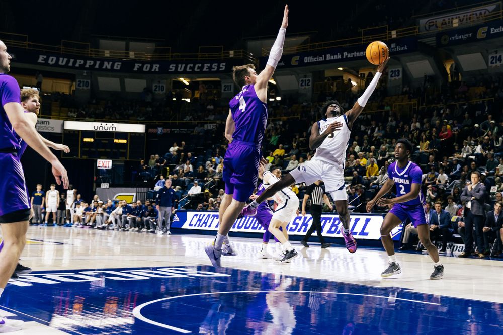 Basketball player leaps for a layup during a game with multiple defenders attempting a block in a blue and white arena.