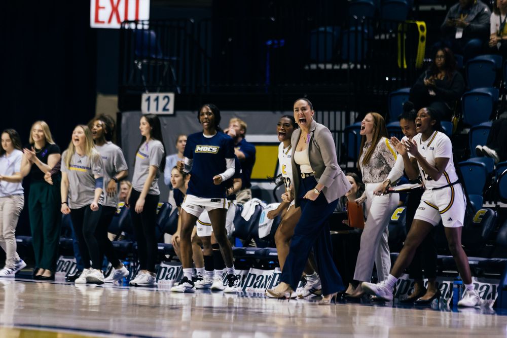 A basketball team and bench players celebrate excitedly during a game, jumping up with enthusiasm at an indoor arena.