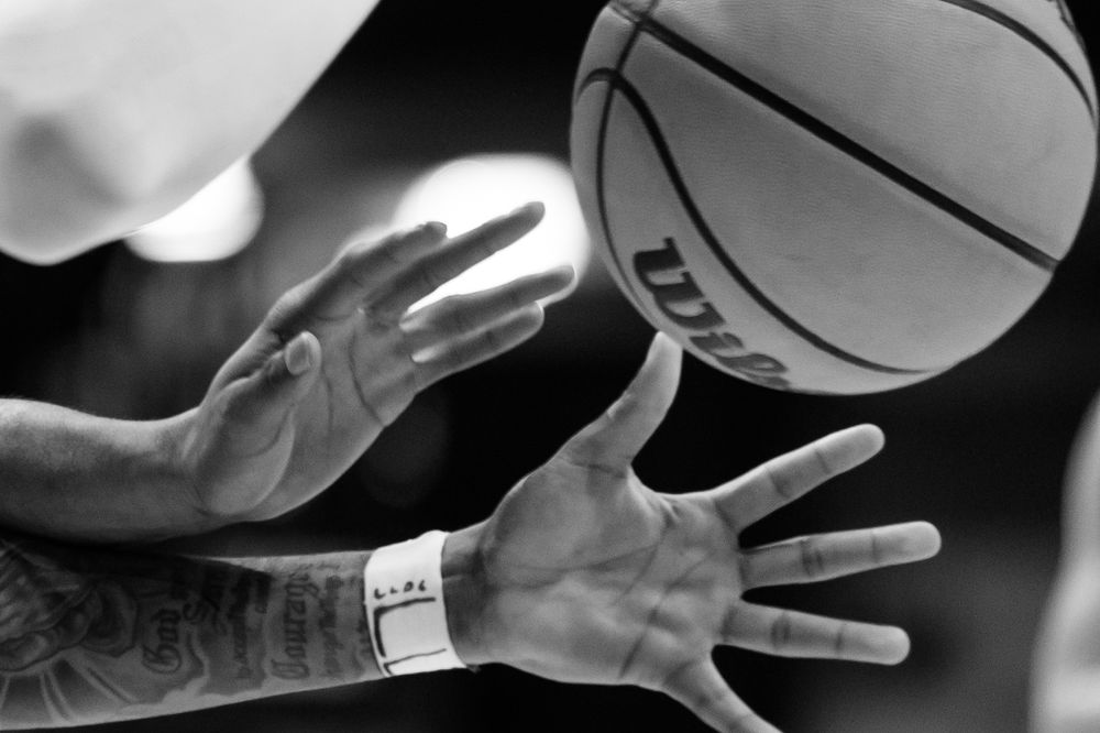 Black and white close-up of tattooed hands reaching for a basketball against dark background.