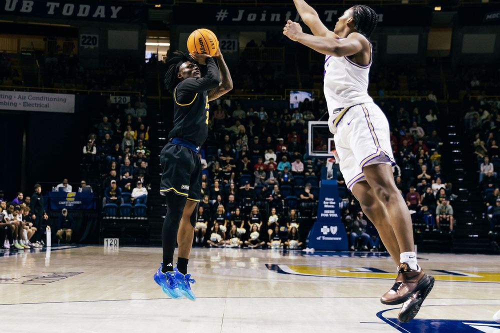 Basketball player in blue jersey takes a jump shot while defender leaps to contest during indoor game.