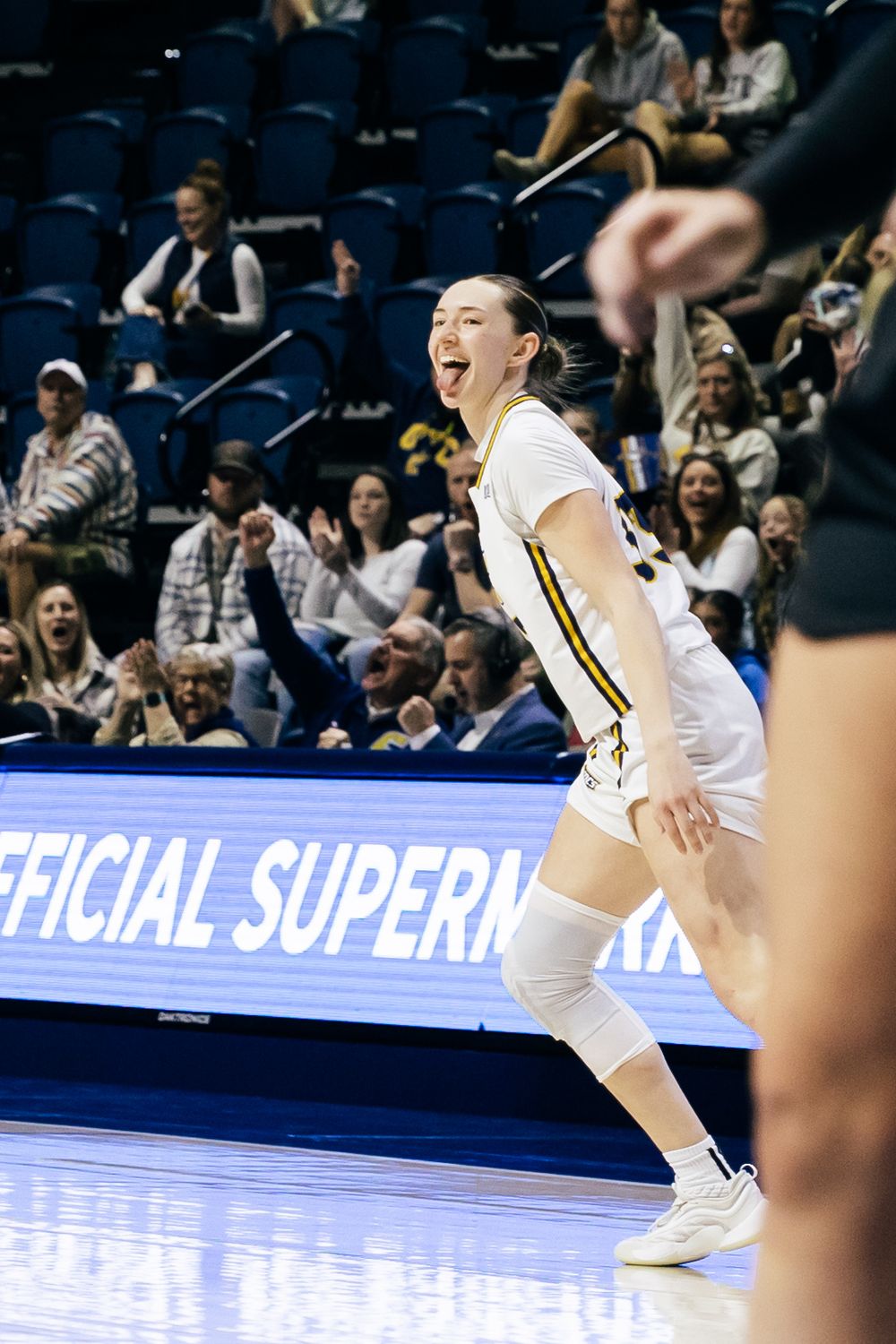 A volleyball player in white uniform stands on a court during a match with stadium seating visible in the background.