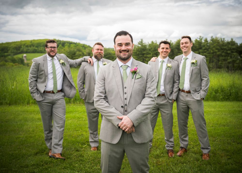 A wedding party in matching gray suits poses together on a lush green lawn under cloudy skies.