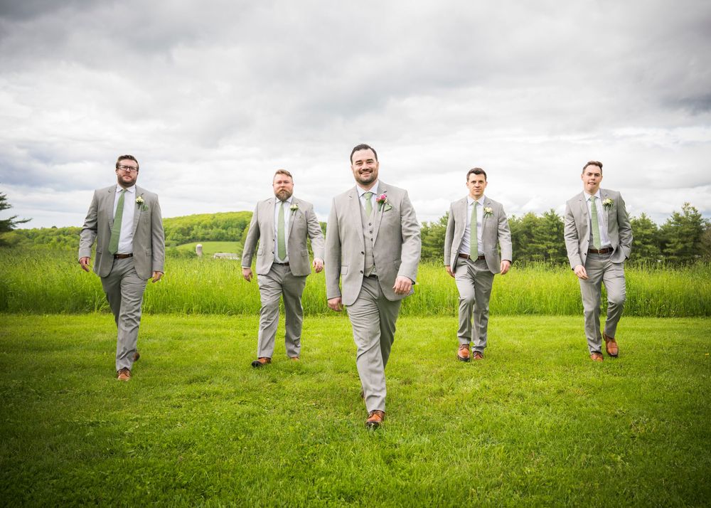 Groomsmen in matching light gray suits walking across a lush green field under cloudy skies.