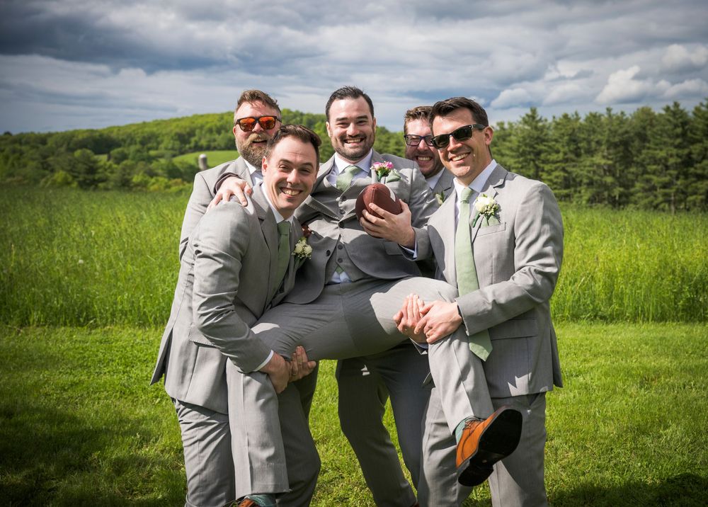 Groomsmen in light gray suits playfully carry the groom while posing on a grassy hill on a cloudy day.