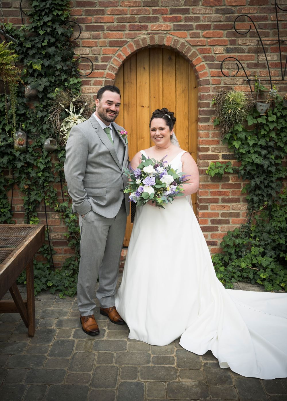 A couple stands before a rustic wooden door framed by ivy and brick in their wedding attire.