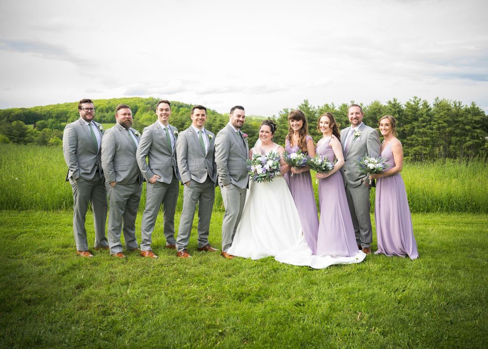 Wedding party in grey suits and lavender dresses pose together on a lush green lawn.