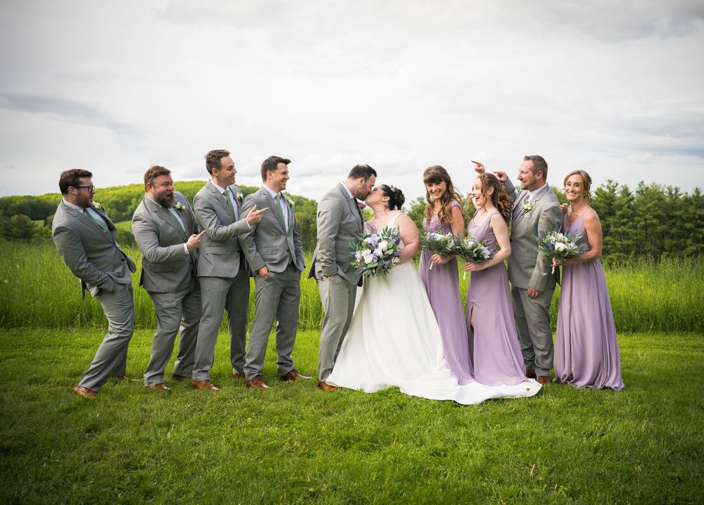 Wedding party in gray suits and lavender dresses posing together on grass field.