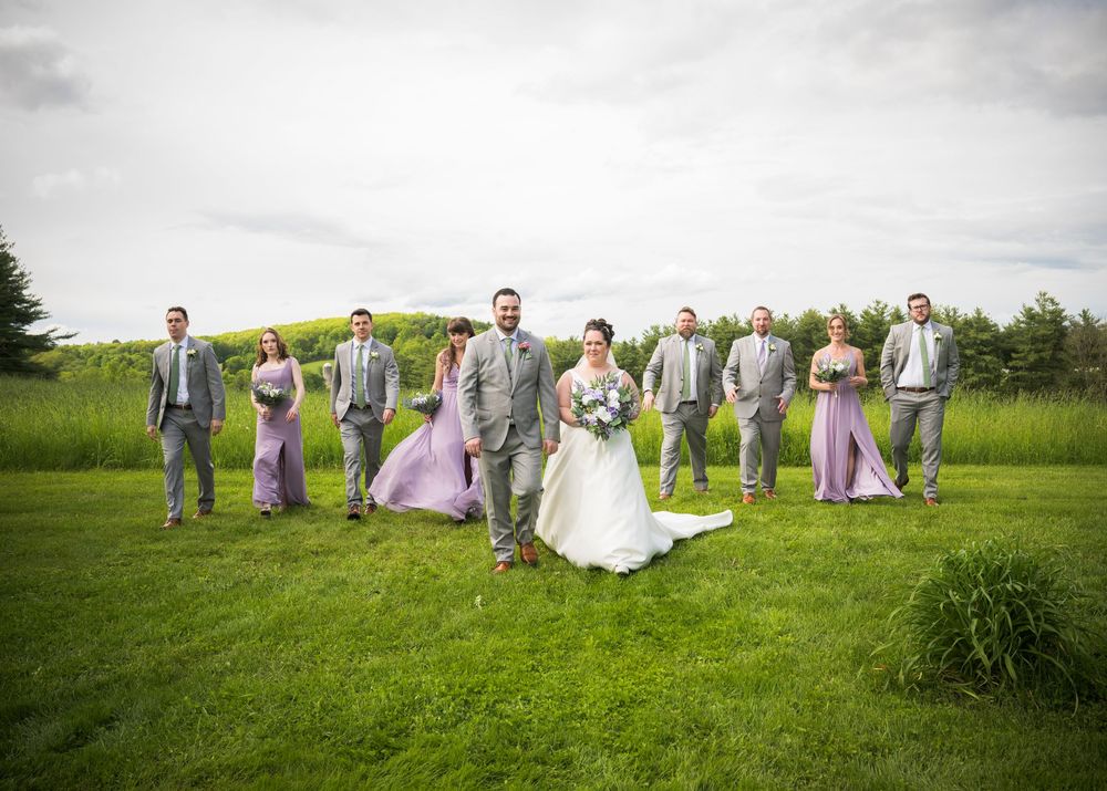 Bridal party in gray suits and purple dresses striking playful poses on grassy field.