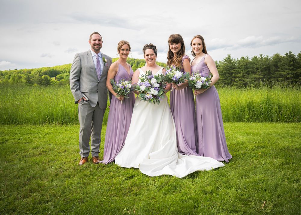 A wedding party poses outdoors with bridesmaids in flowing lavender gowns against a scenic countryside backdrop.