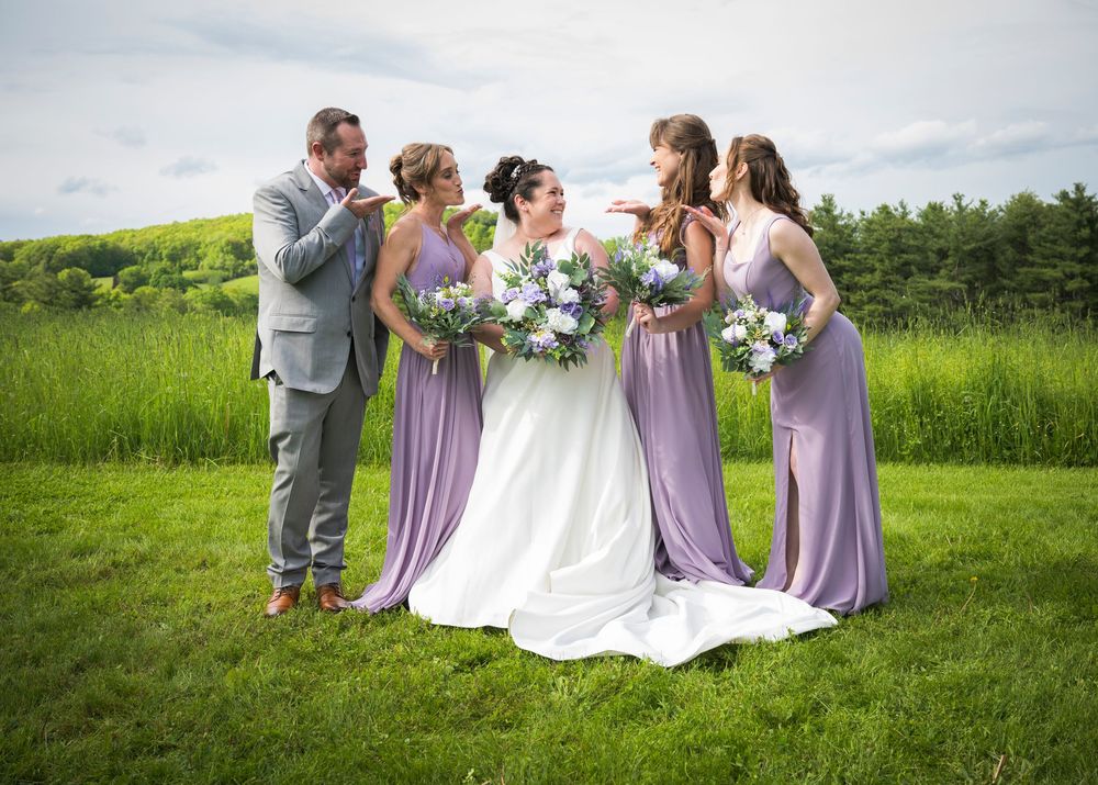 Wedding party in purple dresses and grey suits posing together in a green meadow.