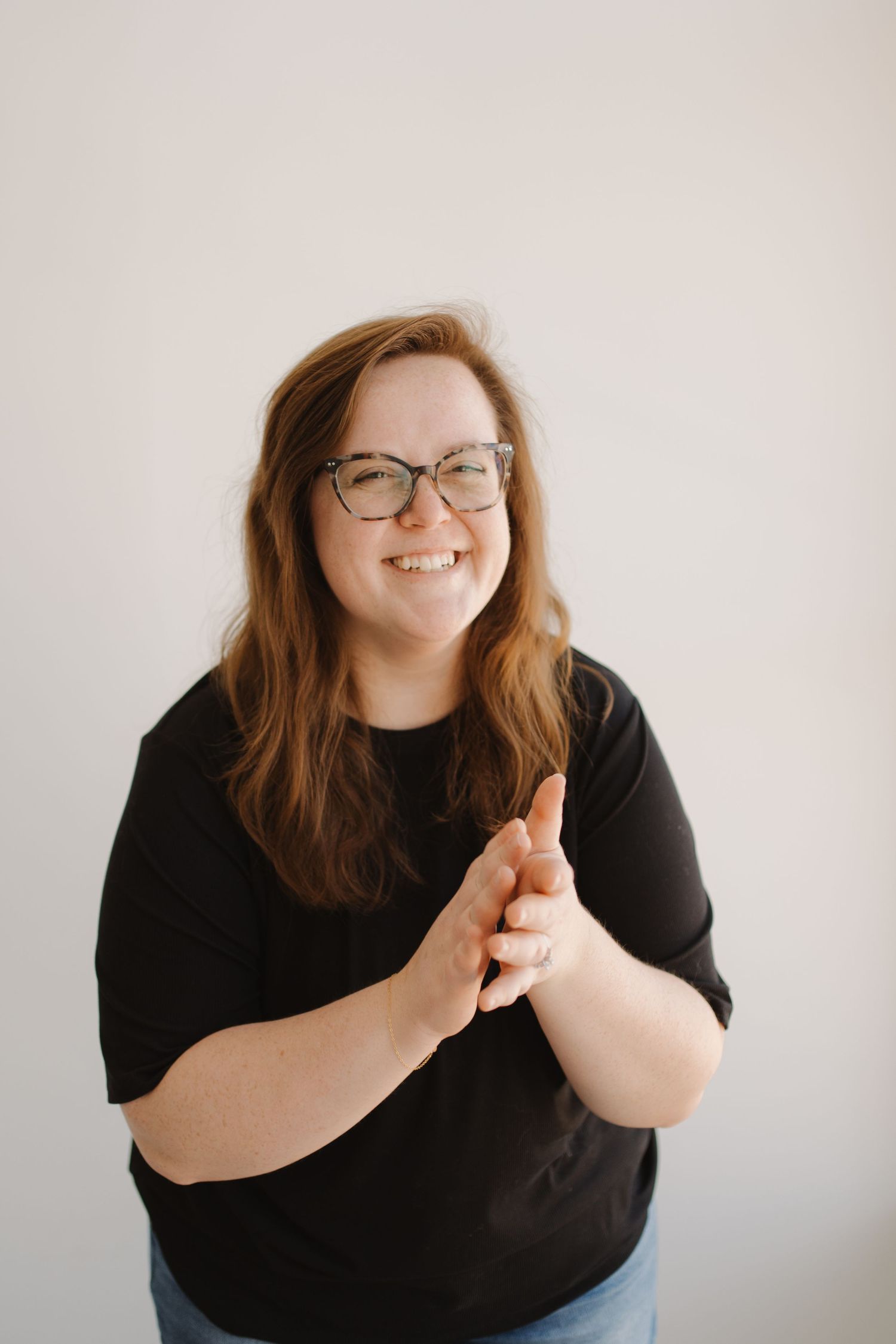 A person wearing glasses and a black shirt stands against a light background while clapping their hands.