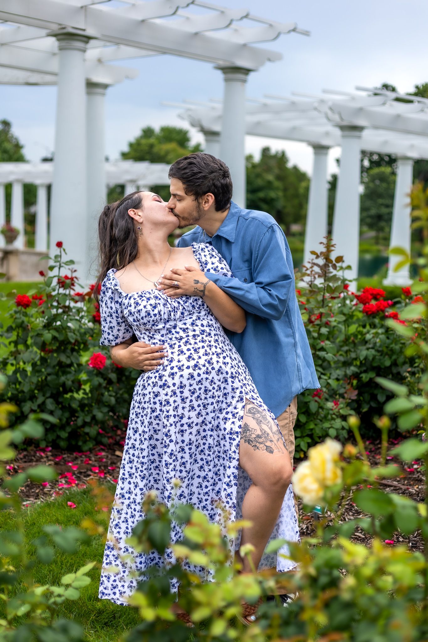 Intimate couple sharing a kiss in a garden with white pergola and red roses.