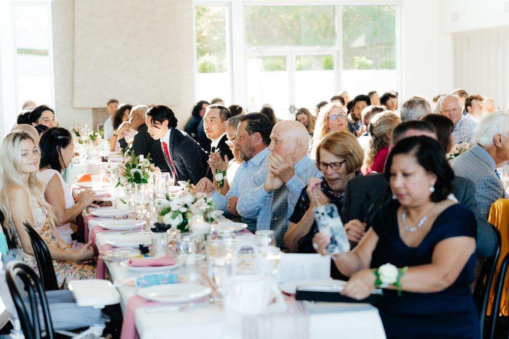 Wedding guests seated at long dining tables with elegant place settings and floral centerpieces at a bright indoor reception.