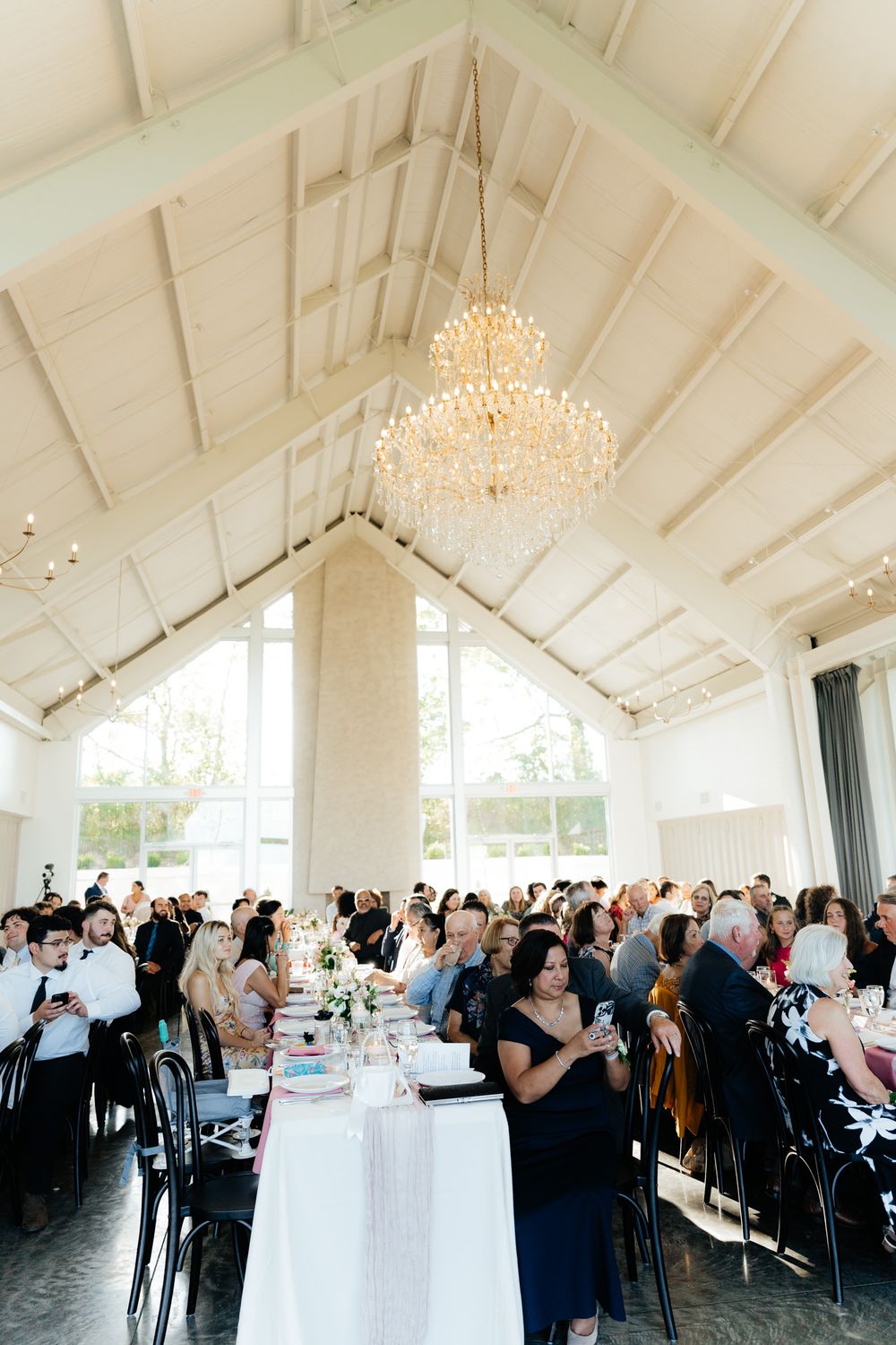A large wedding reception venue with vaulted white ceiling, crystal chandelier, and guests seated at long tables.