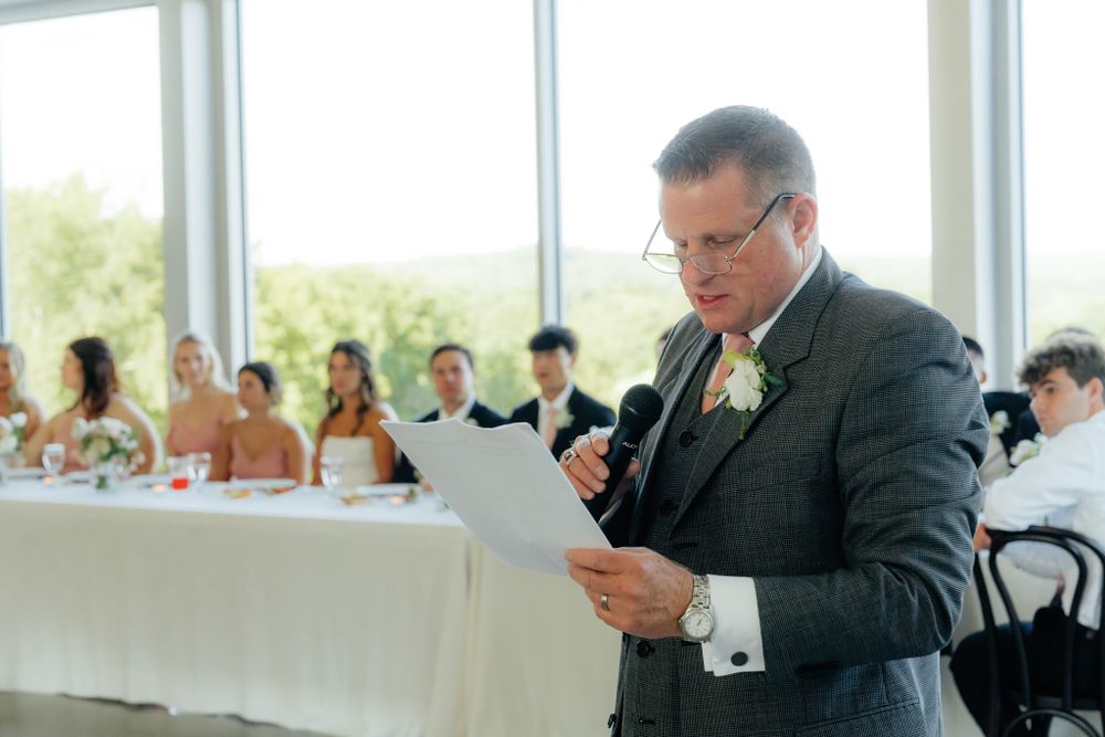 A person in a dark suit gives a speech from a paper while standing near a head table at a formal event.