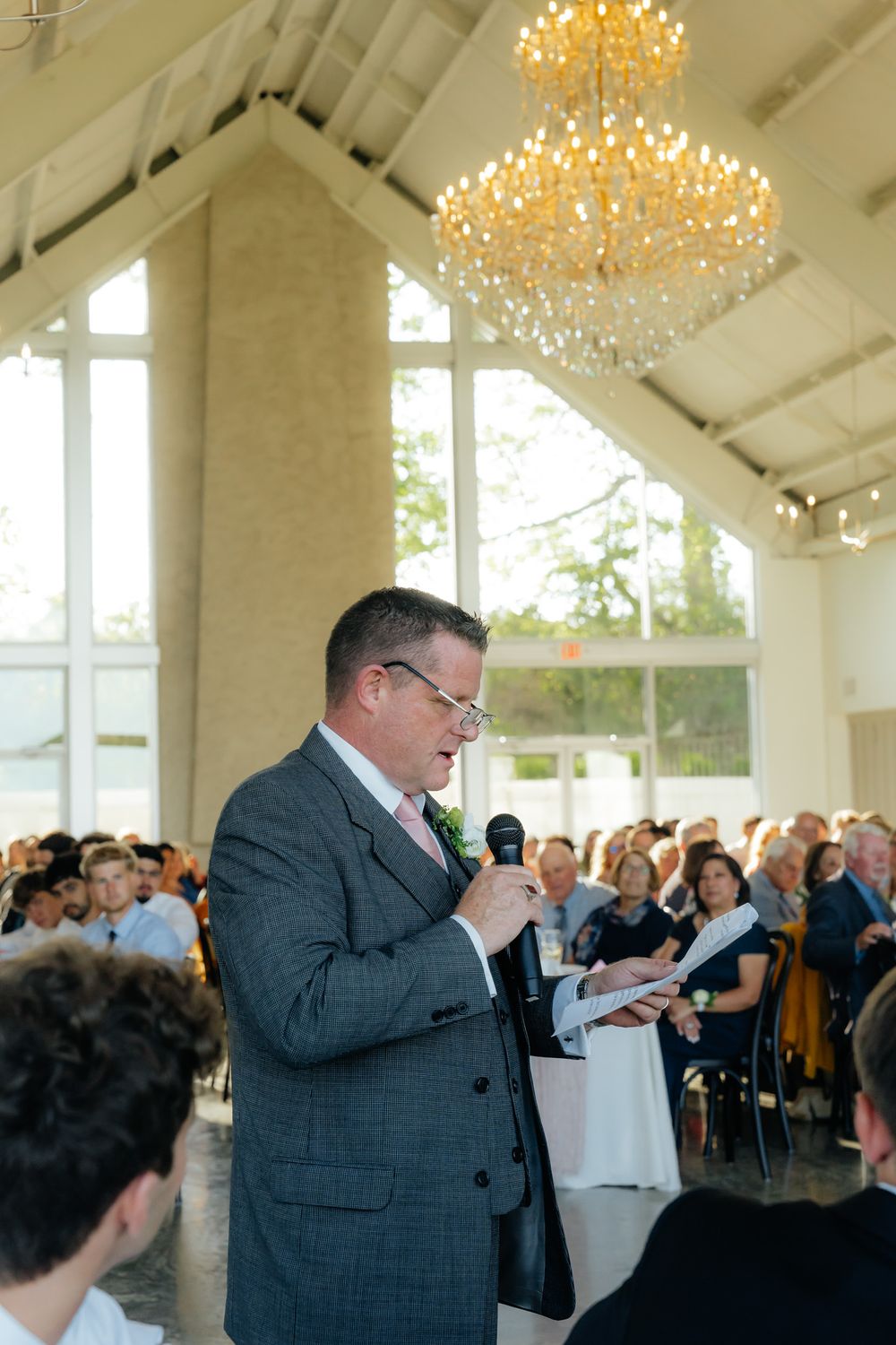Speaker addresses guests in an elegant white venue with crystal chandelier during a special event.