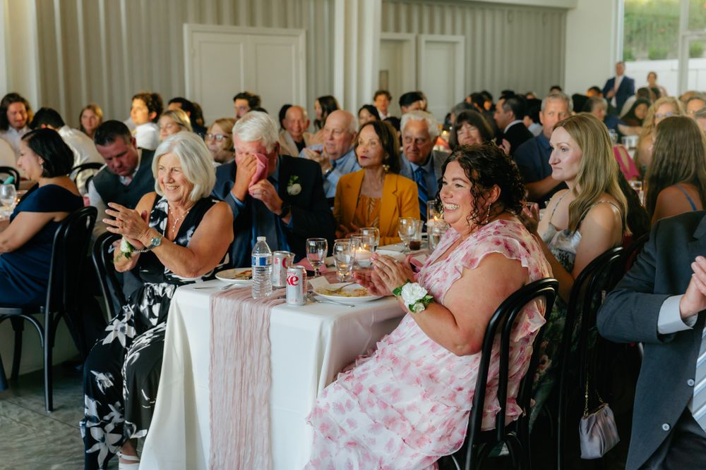 Wedding reception guests enjoy a lively celebration while seated at decorated round tables in a bright indoor venue.