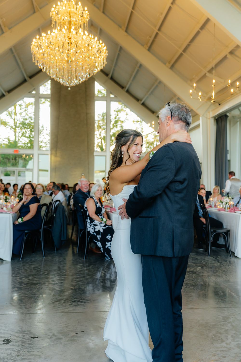 A couple shares their first dance in an elegant venue with a crystal chandelier and wedding reception tables in the background.