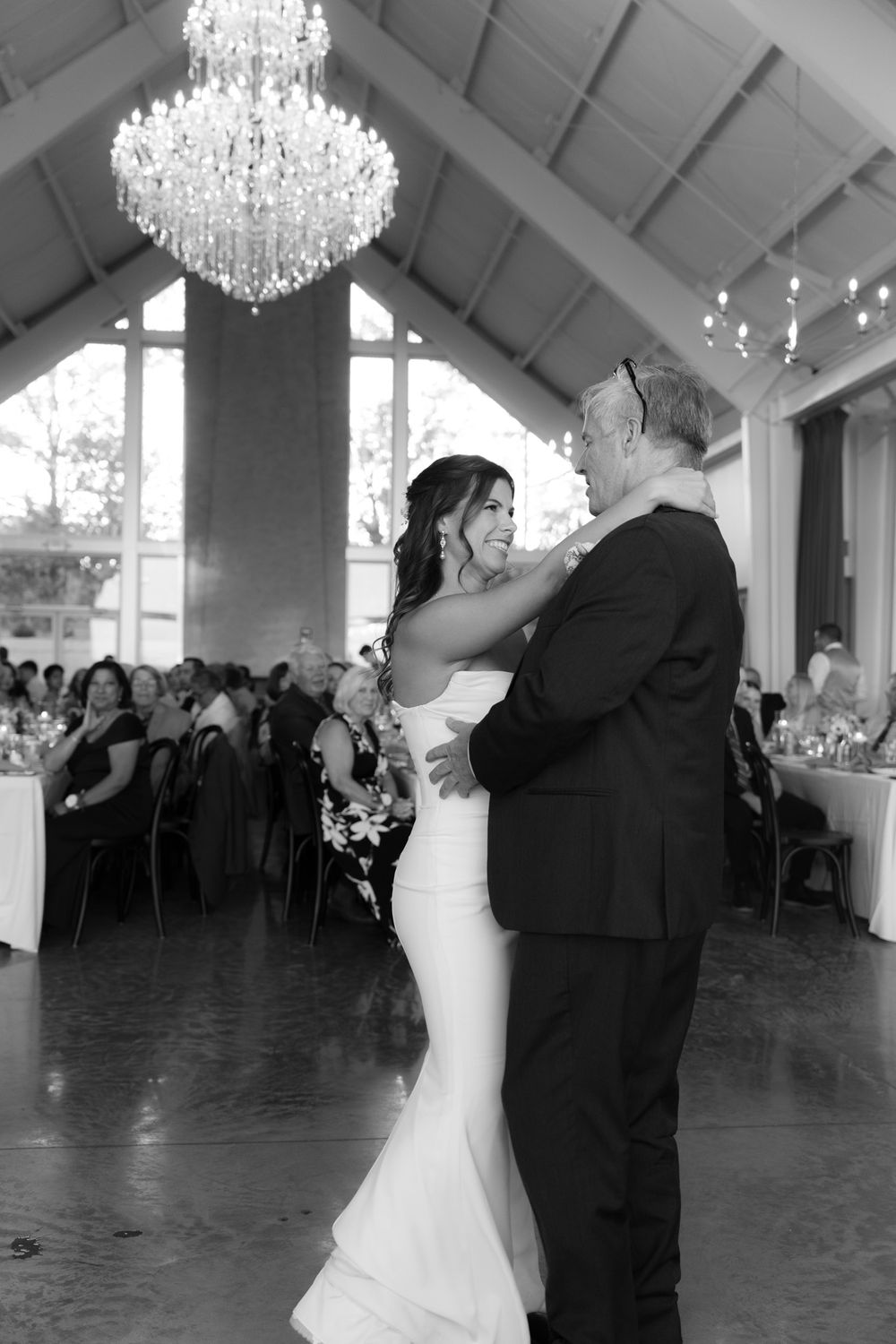 A romantic black and white dance photo of newlyweds beneath a sparkling chandelier at their wedding reception.