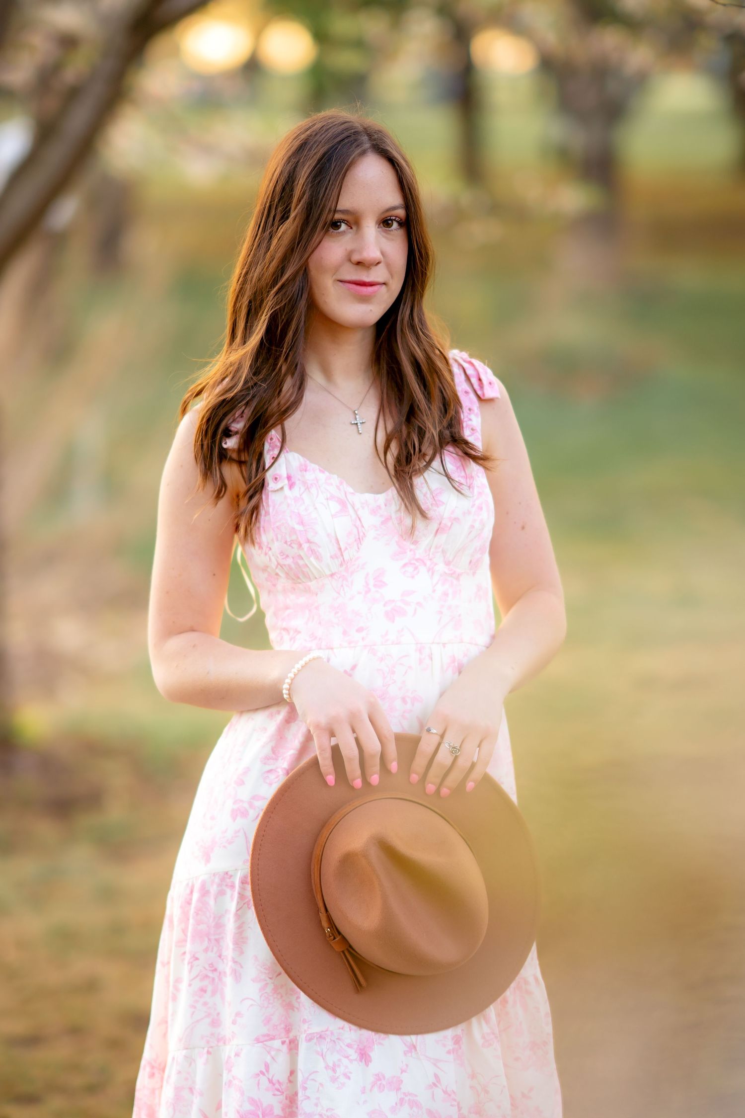 A brown fedora hat is held by someone wearing a floral pink and white sundress in an outdoor setting during golden hour.