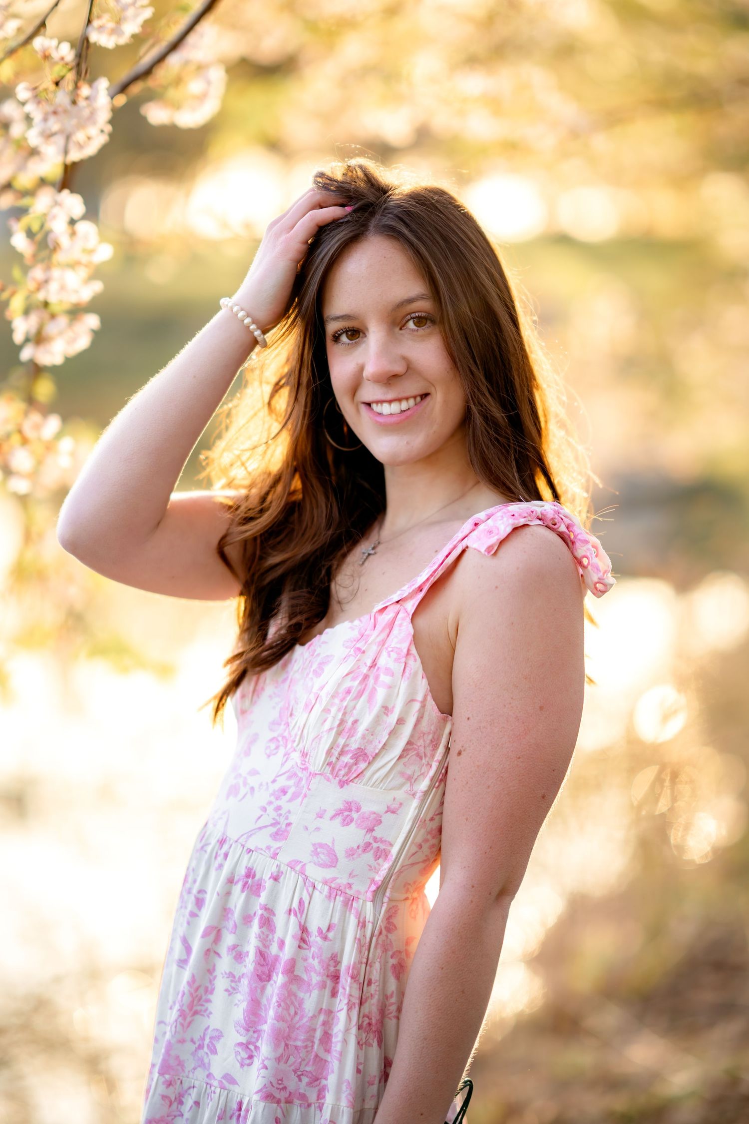 A springtime portrait showing a woman in a pink floral dress posing near blossoming cherry branches in warm golden light.