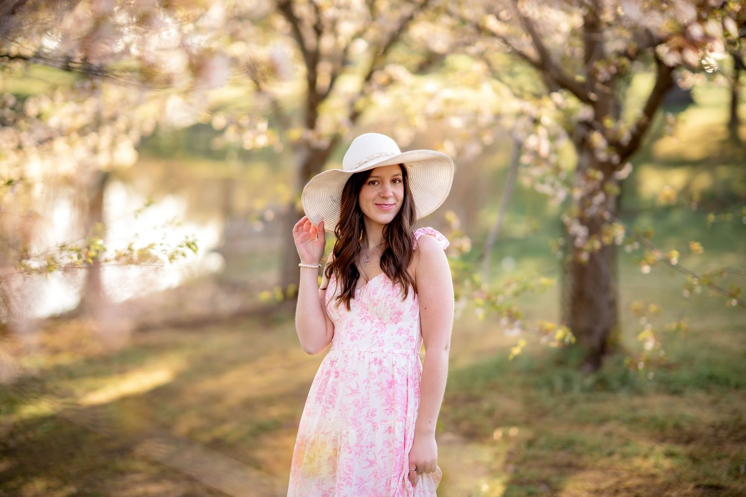 A person in a pink floral dress and sun hat poses beneath blooming cherry blossom trees in soft spring light.
