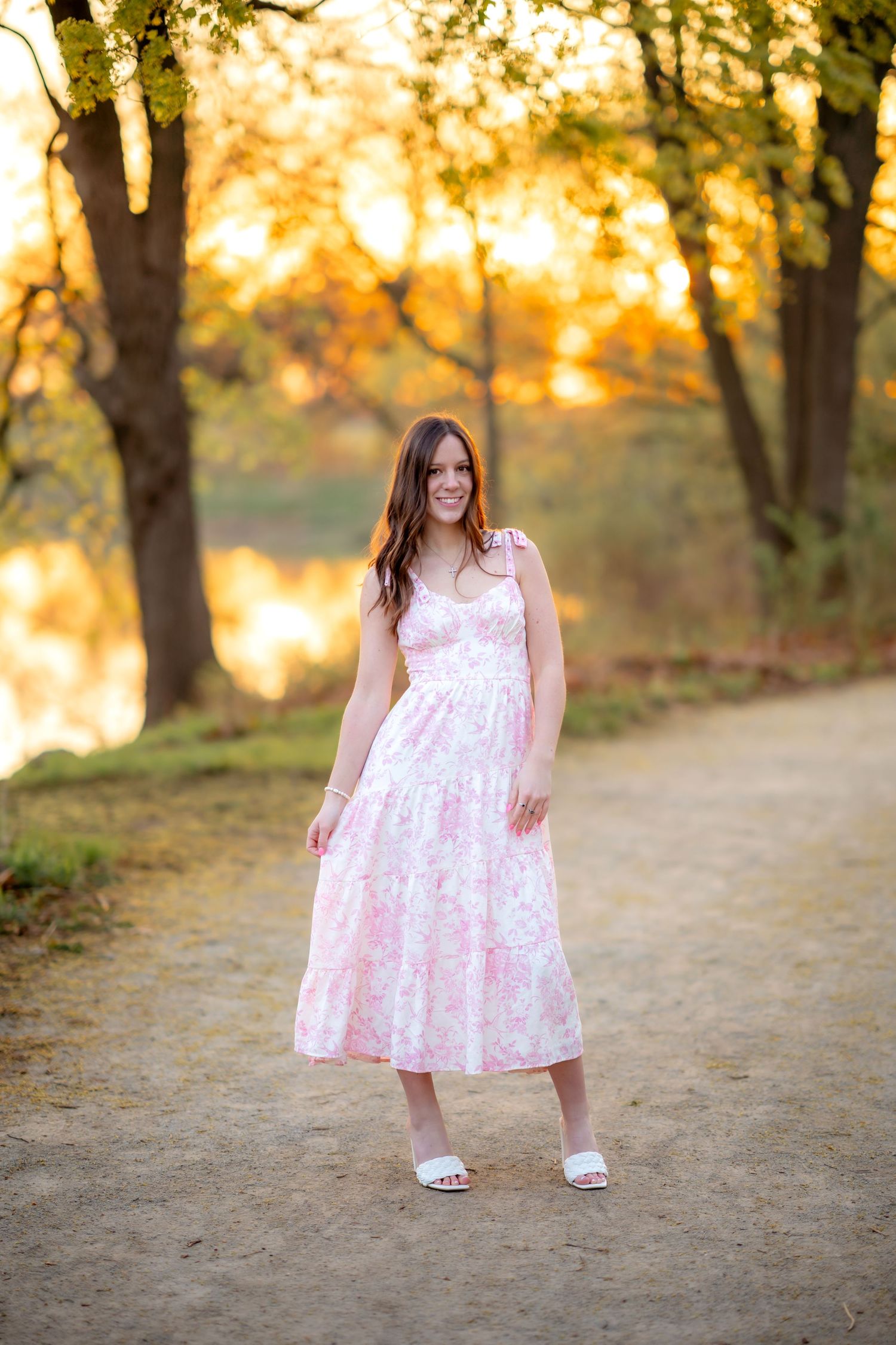 Figure in a pink floral sundress stands on a dirt path at sunset with trees and golden light in the background.