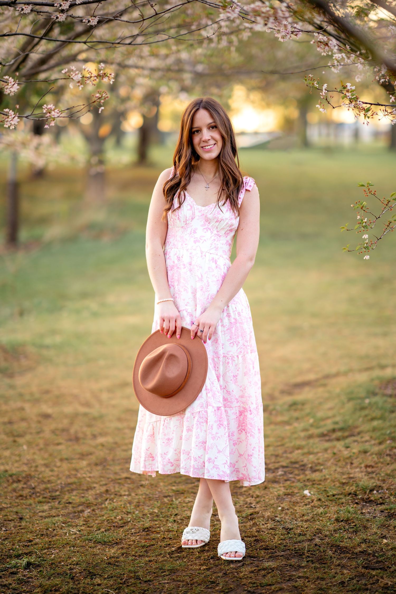 A person in a pink floral sundress and white sandals stands under blooming cherry blossoms holding a brown hat.